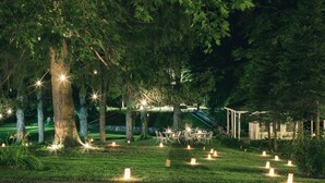 Un área de comedor al aire libre con mesas y sillas bajo árboles, rodeada de velas y un pabellón.