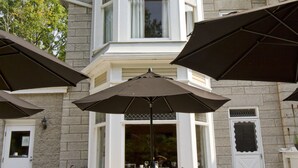 Un patio con mobiliario exterior y sombrillas, un edificio de piedra con ventanal saliente y una puerta con panel decorativo.