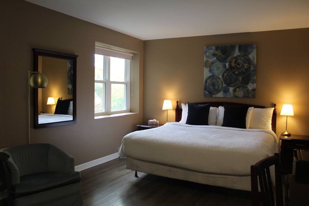 A bedroom with a large bed, two bedside lamps, a chair, a nightstand, a mirror, and a window.