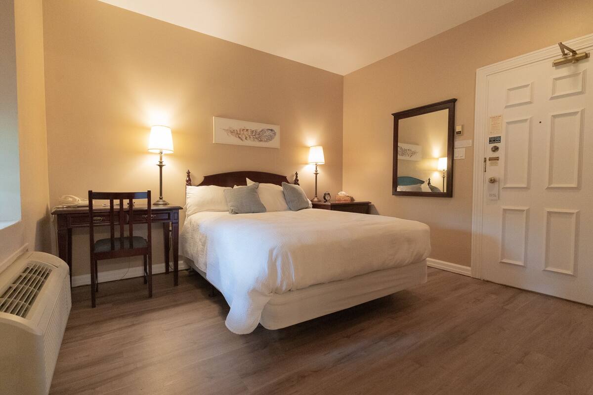 A hotel room with a large bed, two bedside tables with lamps, a chair, a desk, and a door.