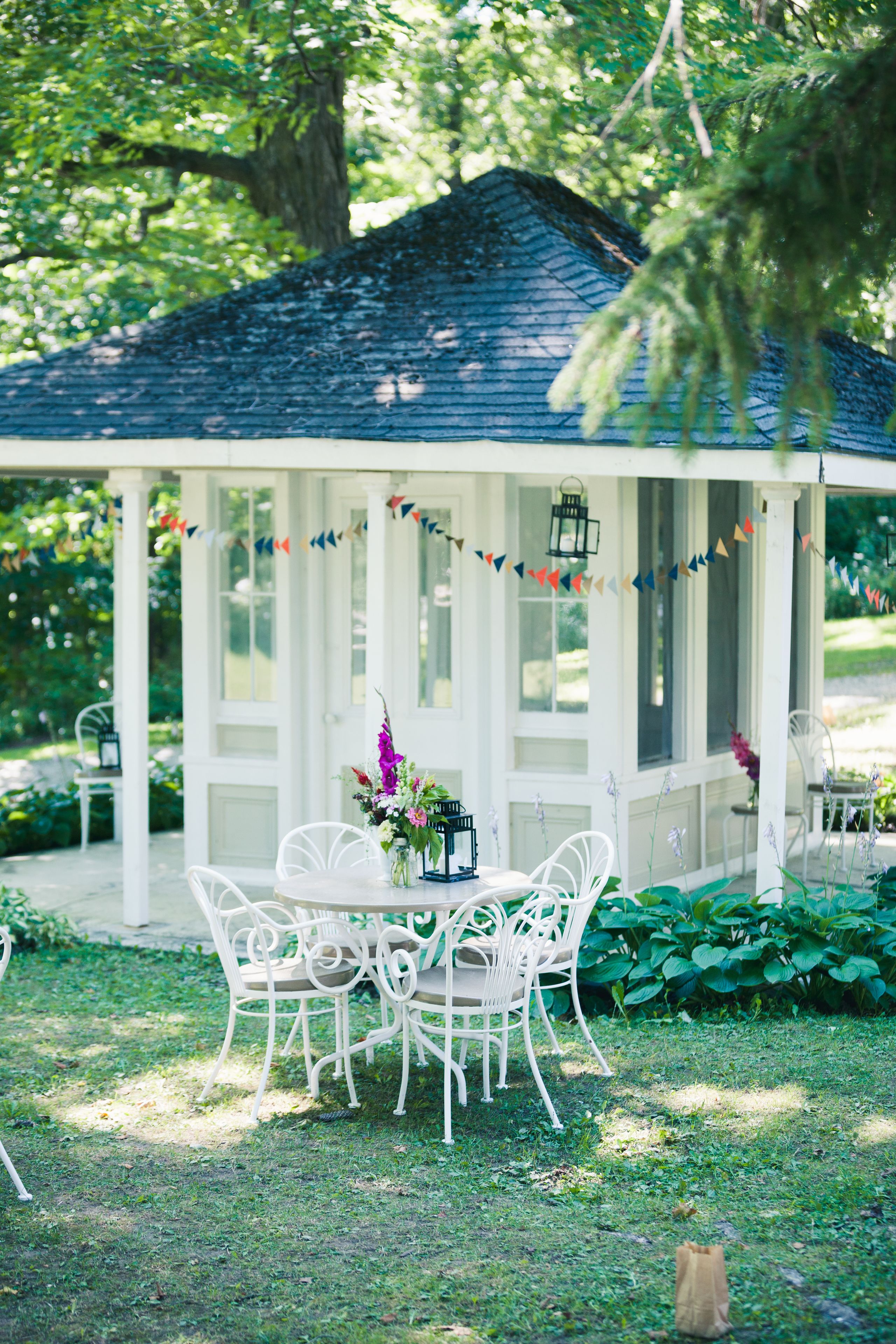 Um gazebo branco com teto azul, rodeado por vegetação e cadeiras brancas.