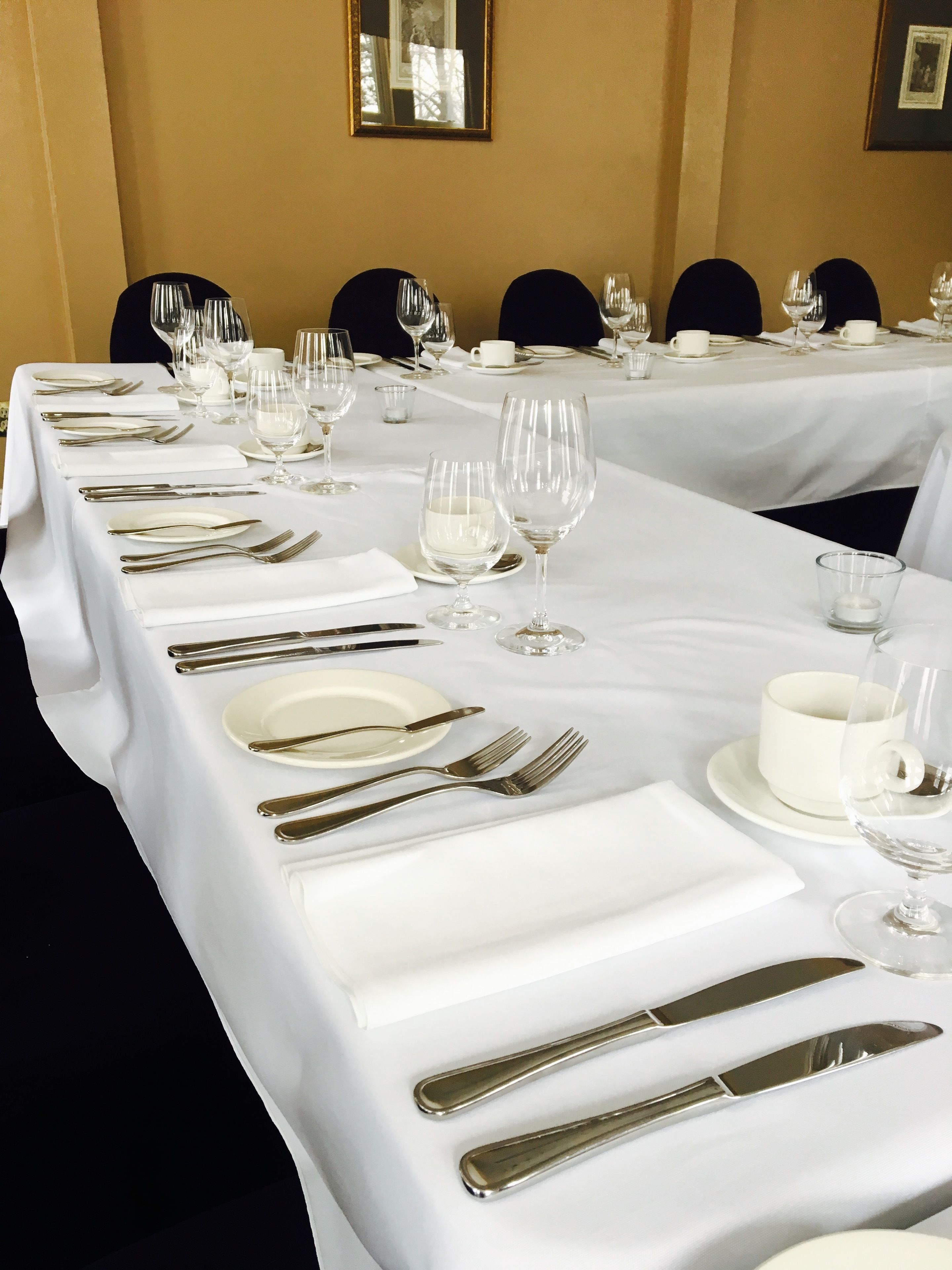 Una larga mesa de comedor dispuesta con manteles blancos, cubiertos y copas de vino, lo que sugiere un espacio para comidas formales.