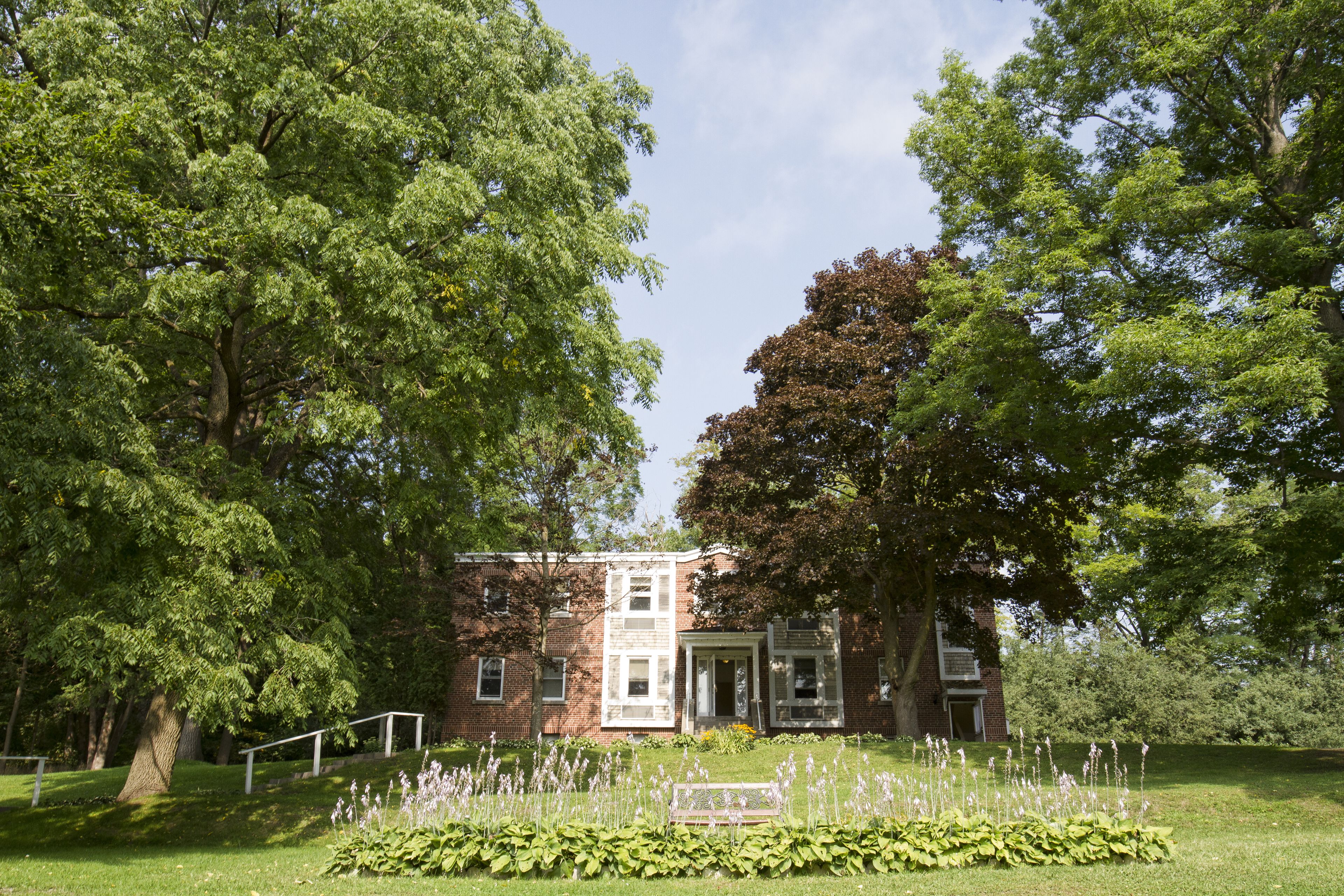 A historic building with a brick facade, large windows, and a well-maintained garden with a bench and flowering plants.