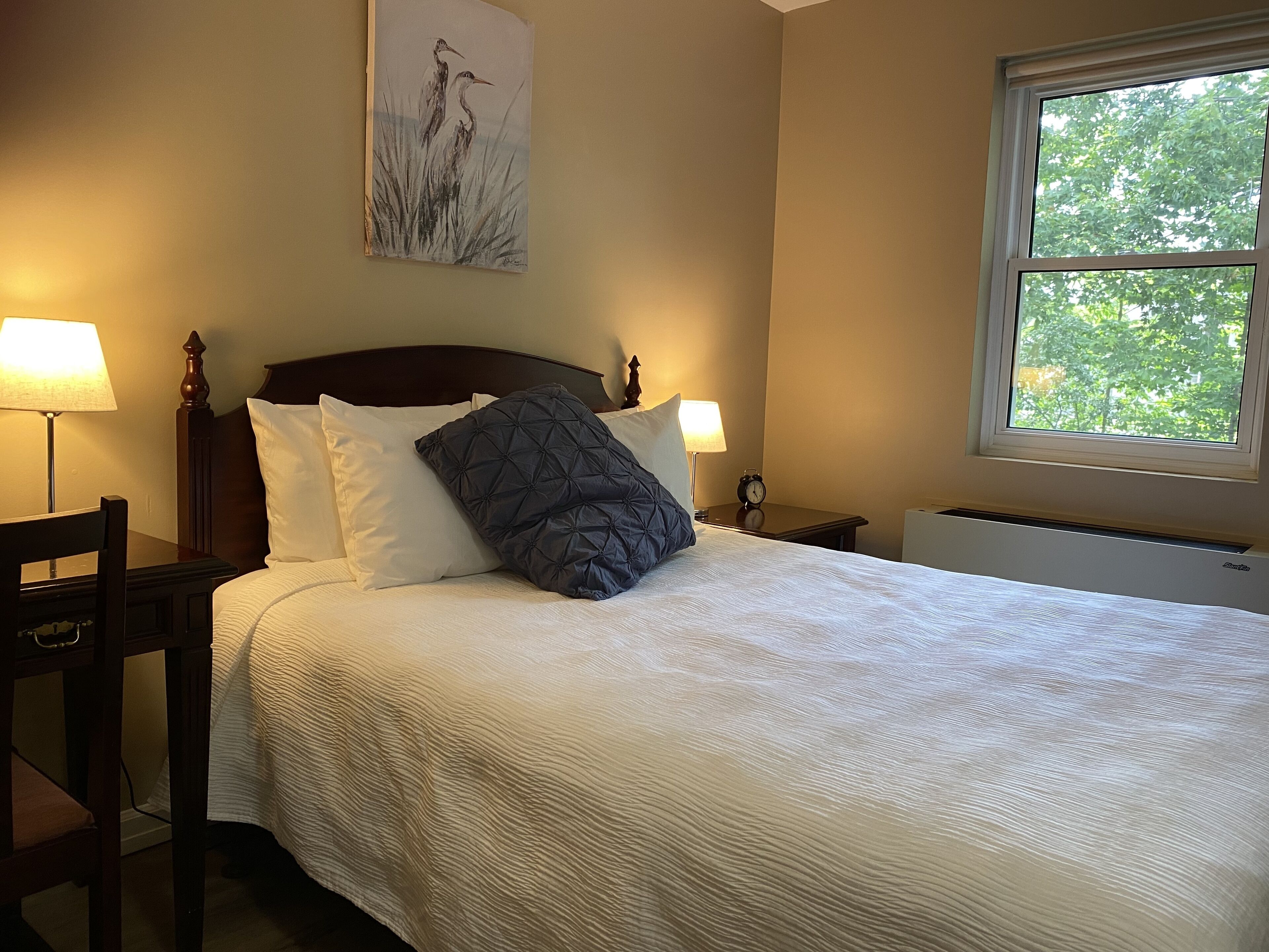 A neatly made bed with white and dark blue pillows, a wooden headboard, and a painting above it. There are two bedside lamps, a clock, and a window with a view of trees.