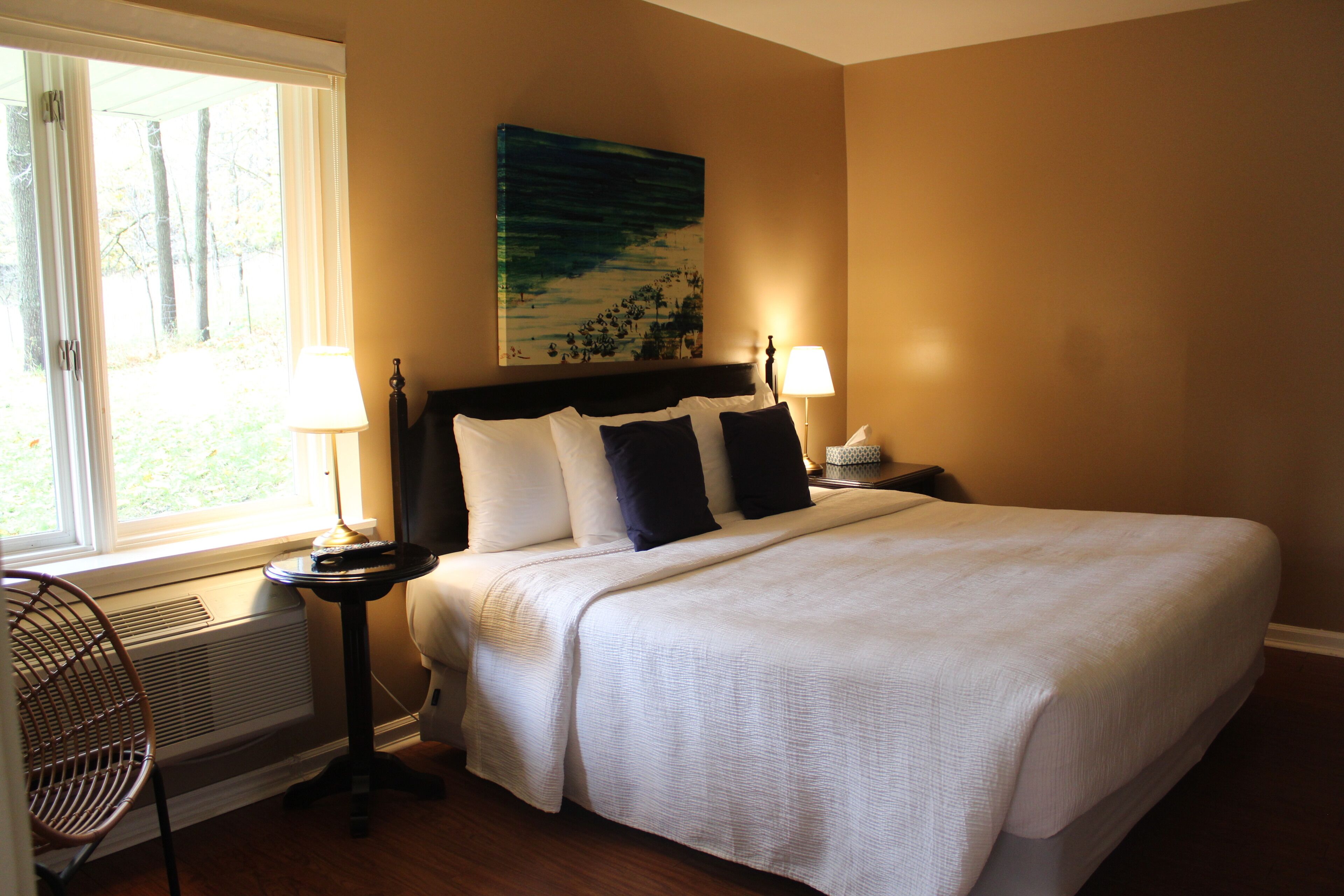 A neatly made bed with white bedding and dark pillows, a bedside table with a lamp, and a chair by the window.