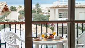 Balcón con mesa dispuesta para el desayuno, que incluye frutas, jugo y pasteles, con vista a una ciudad costera con edificios y palmeras.