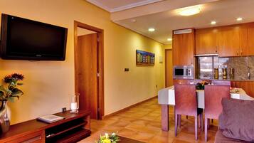 Sala de estar con televisor de pantalla plana, mueble de entretenimiento de madera y una mesa de centro con flores. Se ve un comedor con una mesa y sillas al fondo.