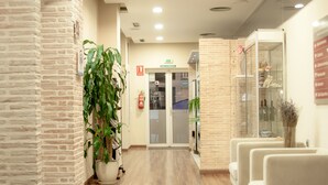 Zona de recepción con pared de ladrillo, planta en maceta, extintor, puerta de vidrio y área de descanso con sillas.