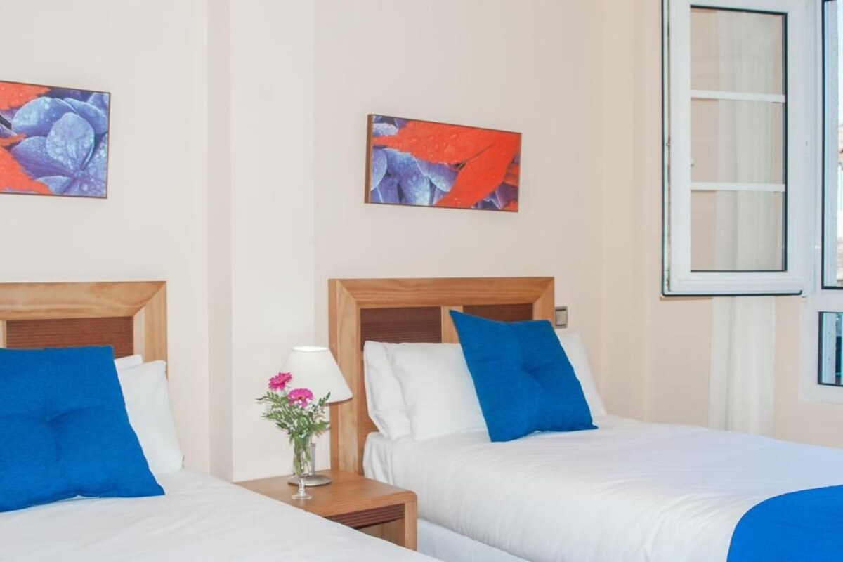 Habitación de hotel con dos camas, cada una con cabecero de madera, ropa de cama blanca y almohadas azules. Entre las camas, un mesita de noche con una lámpara y un jarrón con flores.