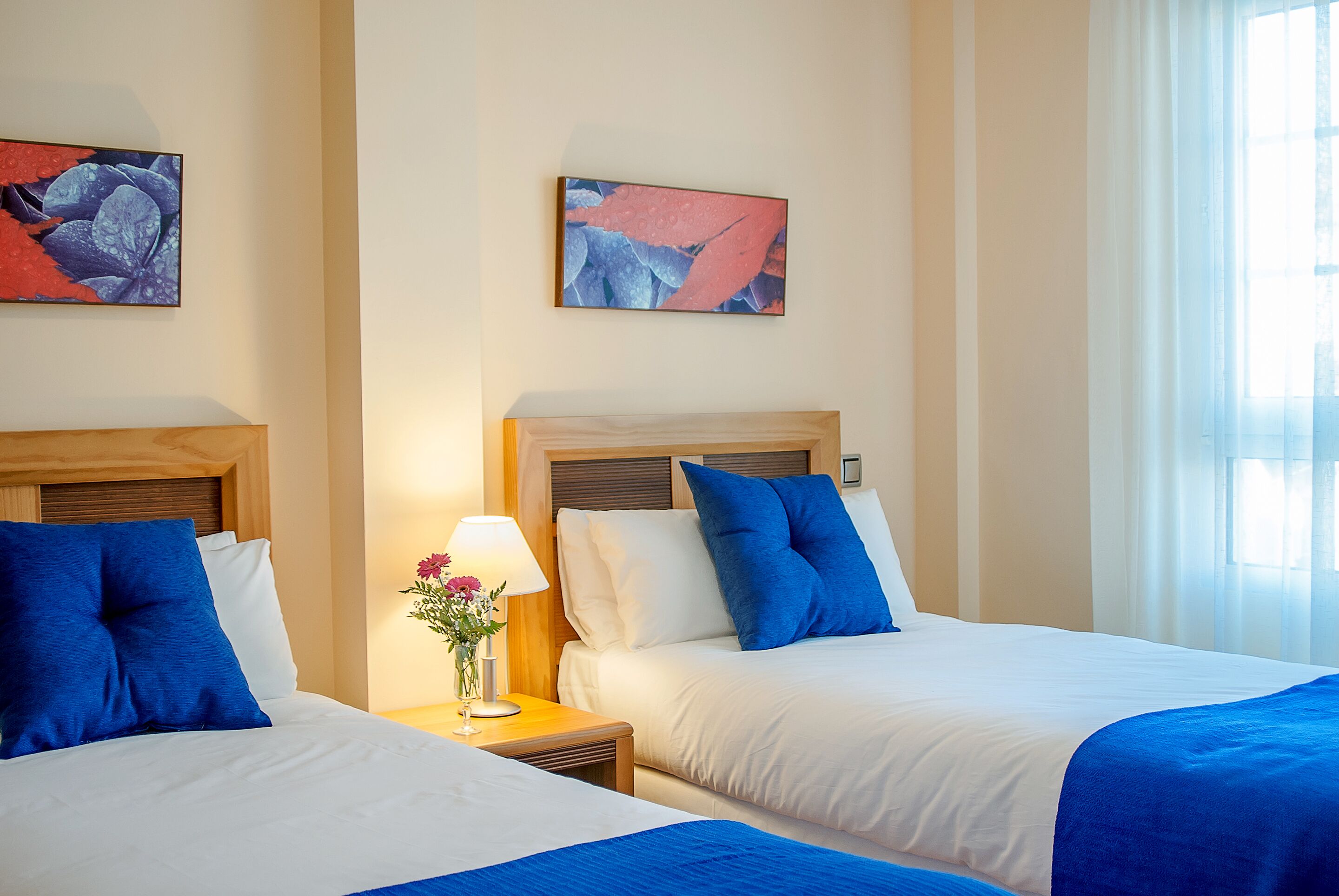 Une chambre d’hôtel avec deux lits, chacun agrémenté de draps bleus et blancs, une table de chevet avec une lampe et des fleurs, et des œuvres d’art accrochées aux murs.
