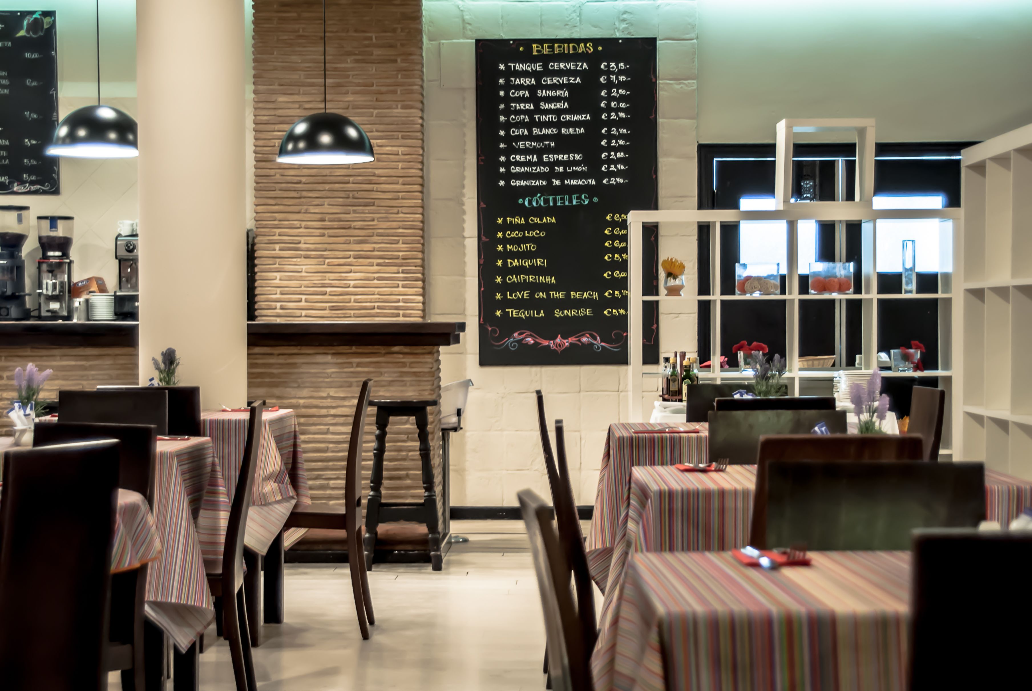 A restaurant with tables covered in striped tablecloths, a menu on the wall, and pendant lighting.