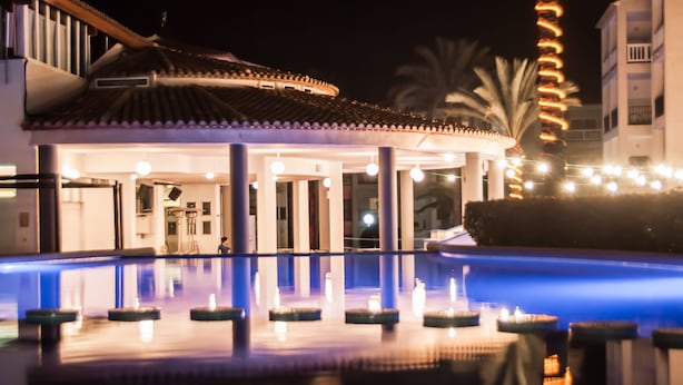 Un área de piscina con fuente de agua, palmeras y un edificio con techo de tejas.