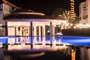 Un área de piscina con fuente de agua, palmeras y un edificio con techo de tejas.