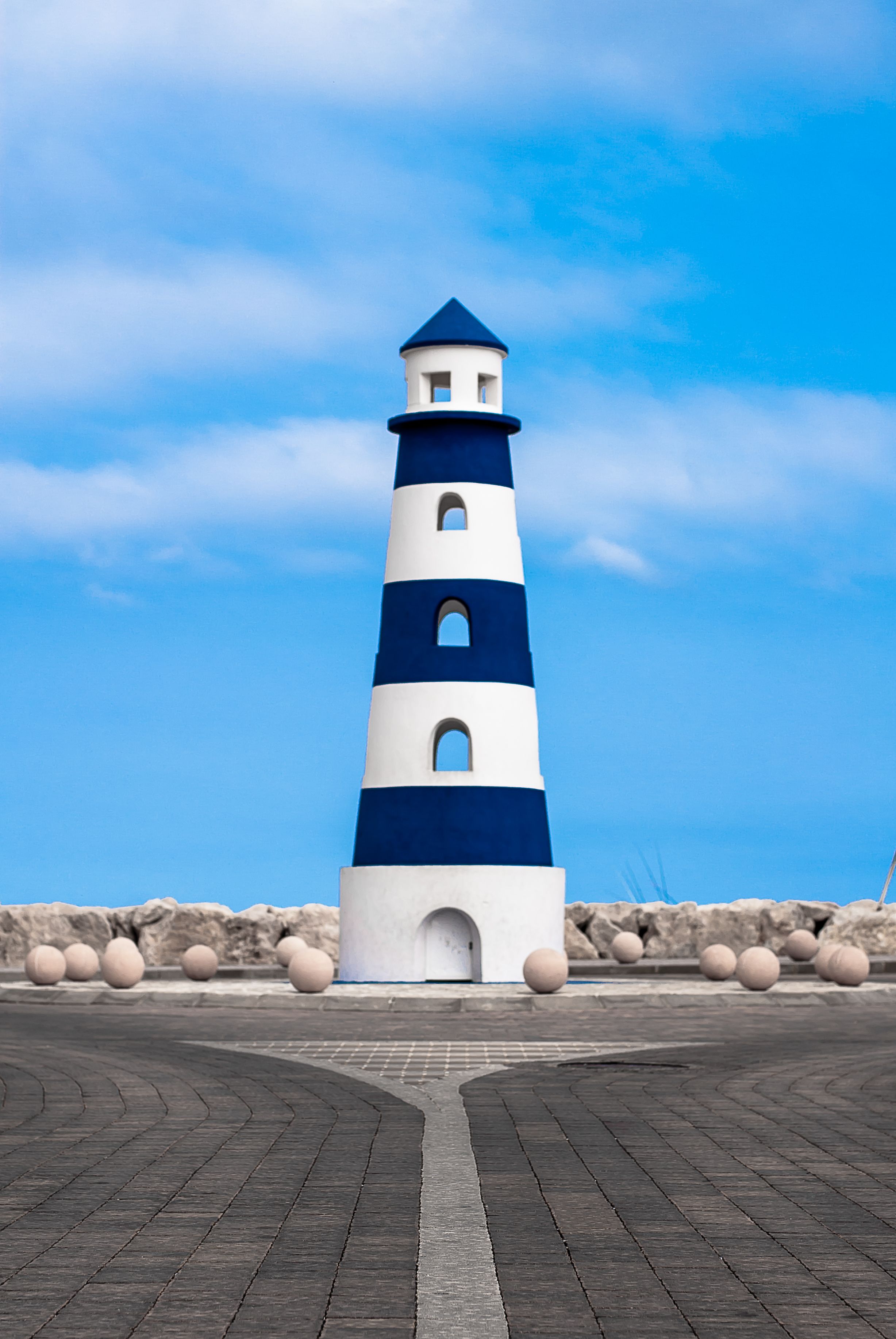 Een vuurtoren met een wit- en blauw kleurenschema, gelegen op een verhard gebied met een heldere lucht op de achtergrond.