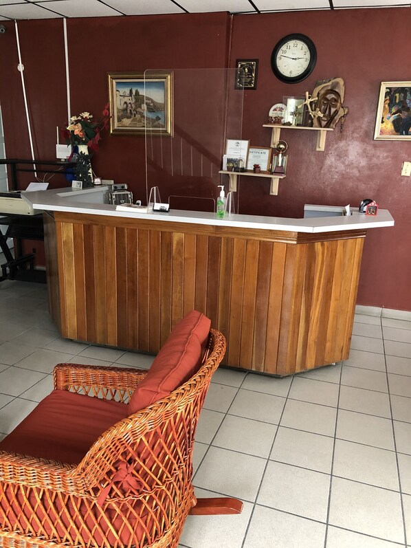 Un área de recepción con mostrador de madera, una silla de mimbre con cojín rojo, un reloj y diversas decoraciones en la pared.