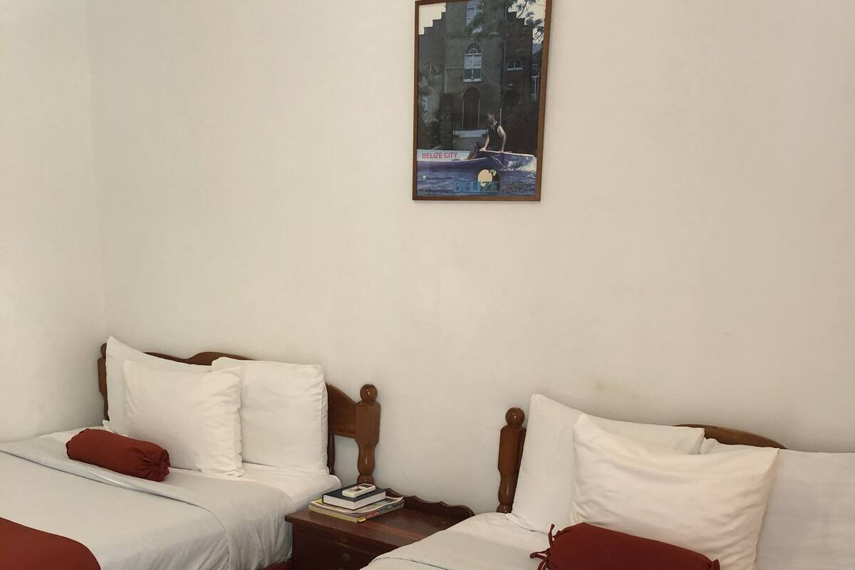Habitación de hotel con dos camas, cada una con ropa blanca y detalles rojos, cabeceros de madera y mesitas de noche con un libro y una lámpara.