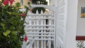Una entrada protegida con valla de piquetes blancos, un camino de grava y un edificio de fachada amarilla al fondo.