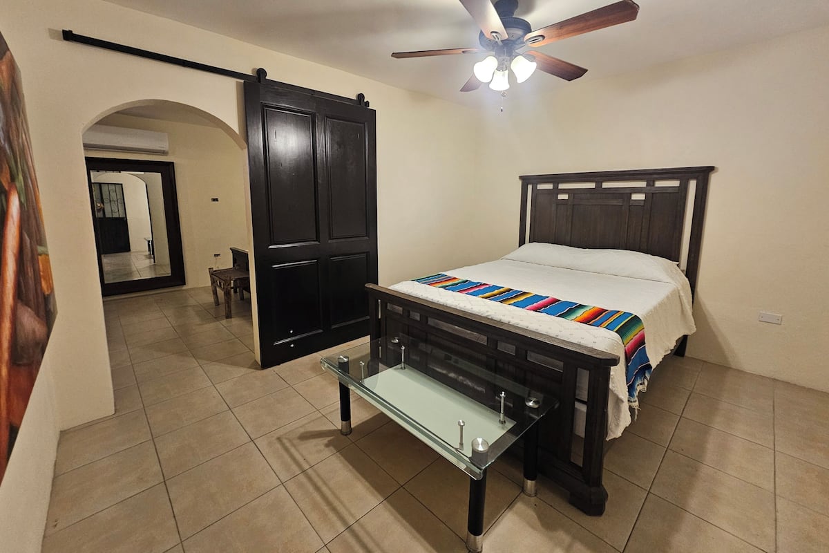 A bedroom with a bed, a glass table, a ceiling fan, and a door leading to another room.