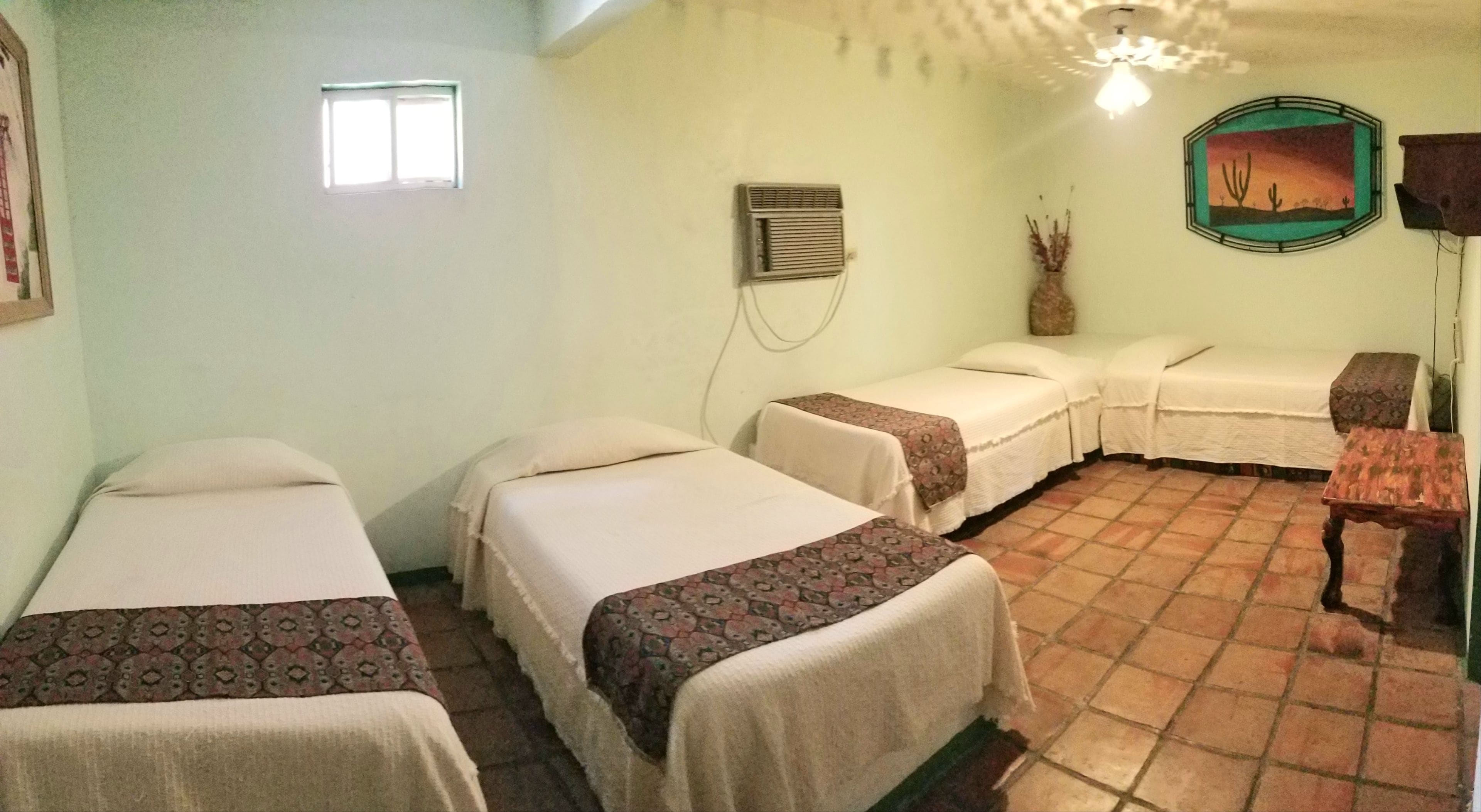 A room with three single beds, a small table, a wall-mounted air conditioning unit, and a desert-themed picture.