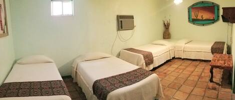 A room with three single beds, a small table, a wall-mounted air conditioning unit, and a desert-themed picture.