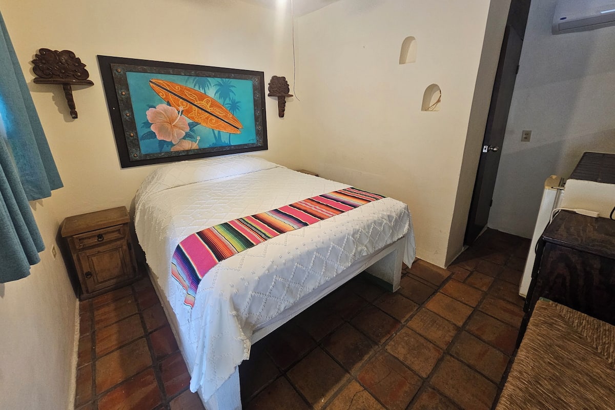 A bedroom with a bed, a small wooden nightstand, a framed picture on the wall, and a door to another room.