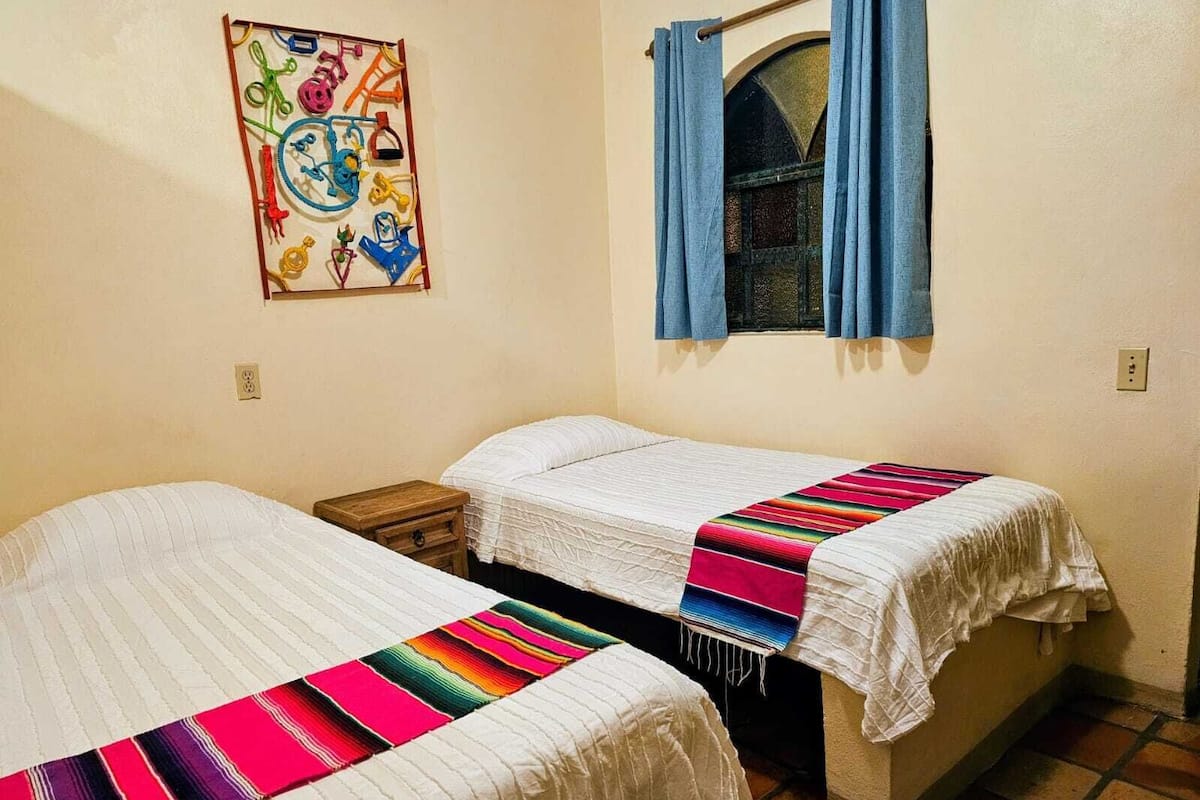 A small, cozy room with two single beds, a wooden nightstand, a window with blue curtains, and a colorful artwork on the wall.