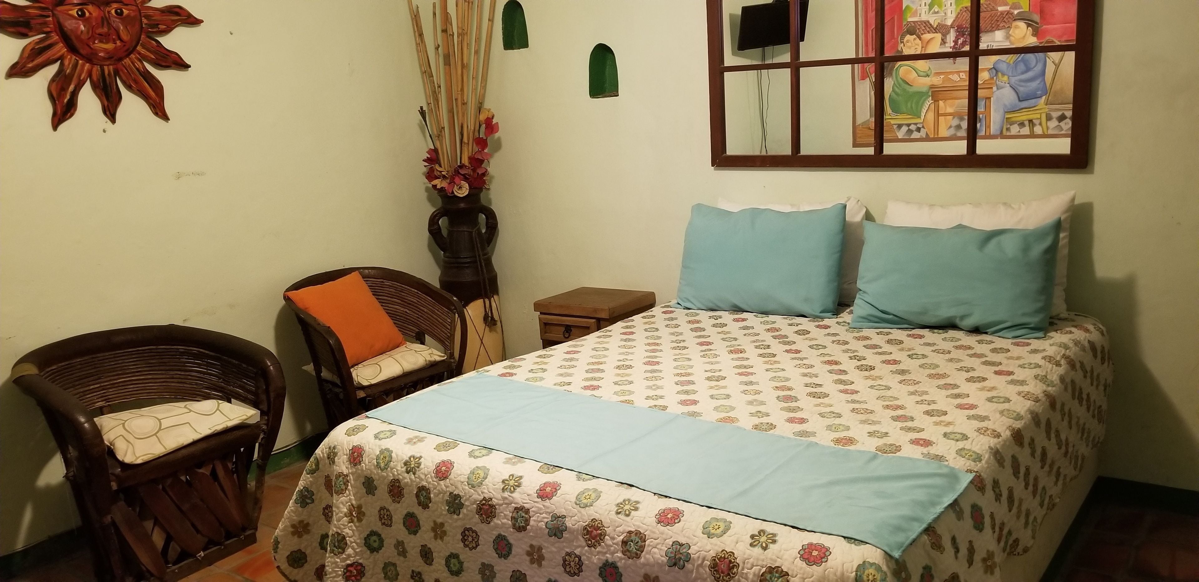 A bedroom with a bed, two pillows, a chair, a small table, a wall-mounted TV, and a decorative sun and flower arrangement.