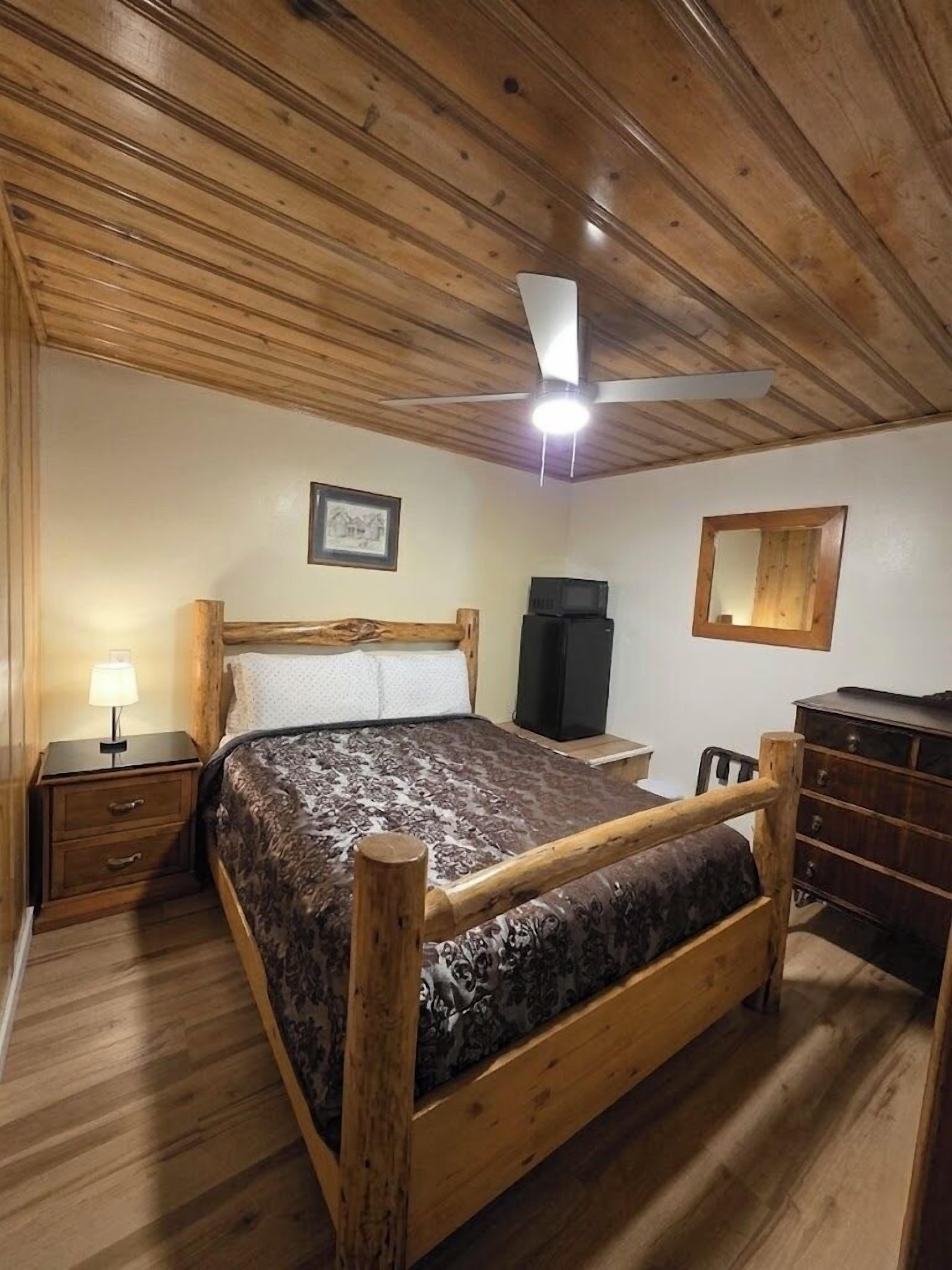 A bedroom with a wooden ceiling, a bed with a headboard, a nightstand, a dresser, a ceiling fan, and a framed picture on the wall.