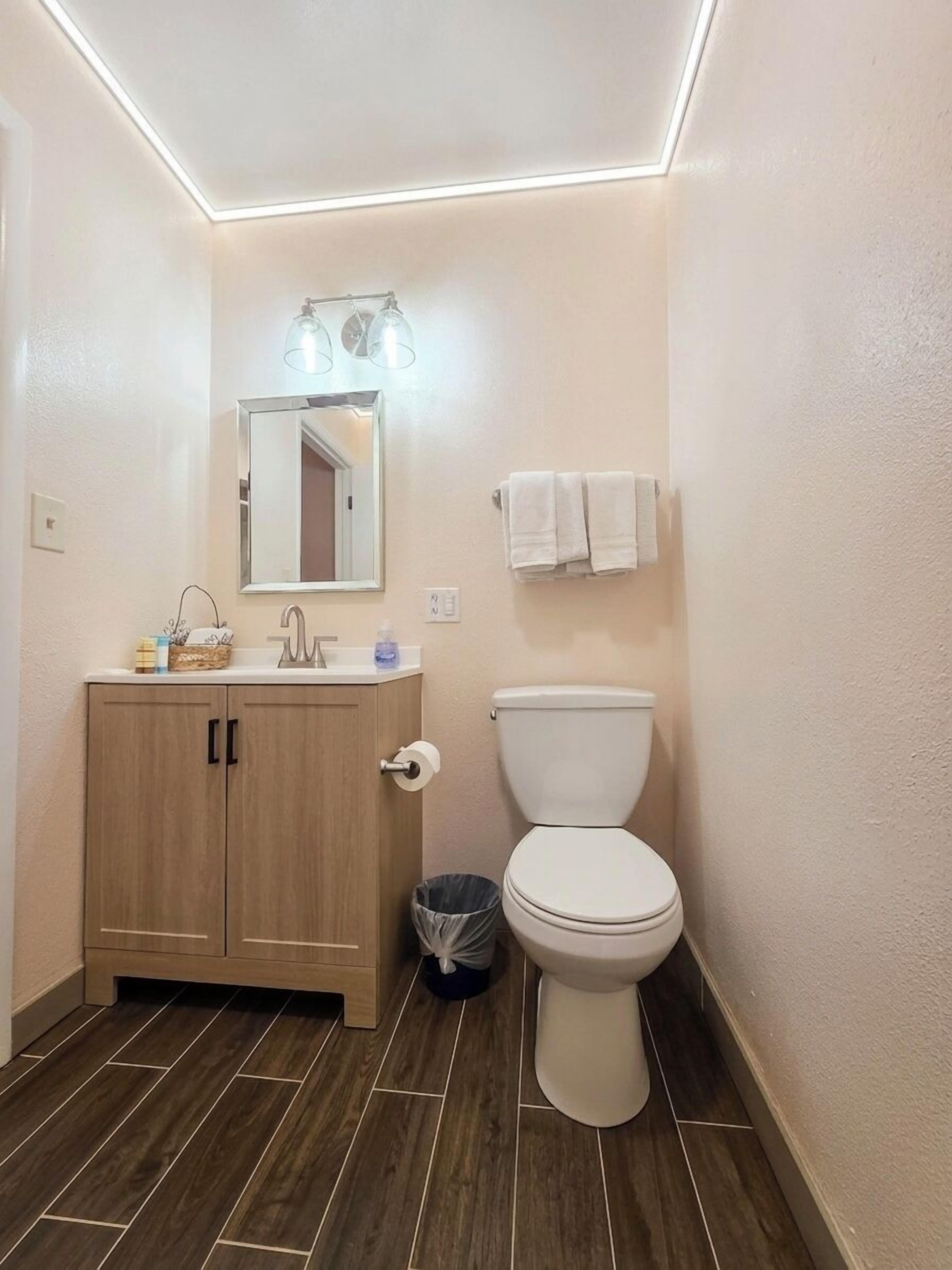 Un baño con un mueble de madera, un inodoro, un espejo y un cubo de basura.