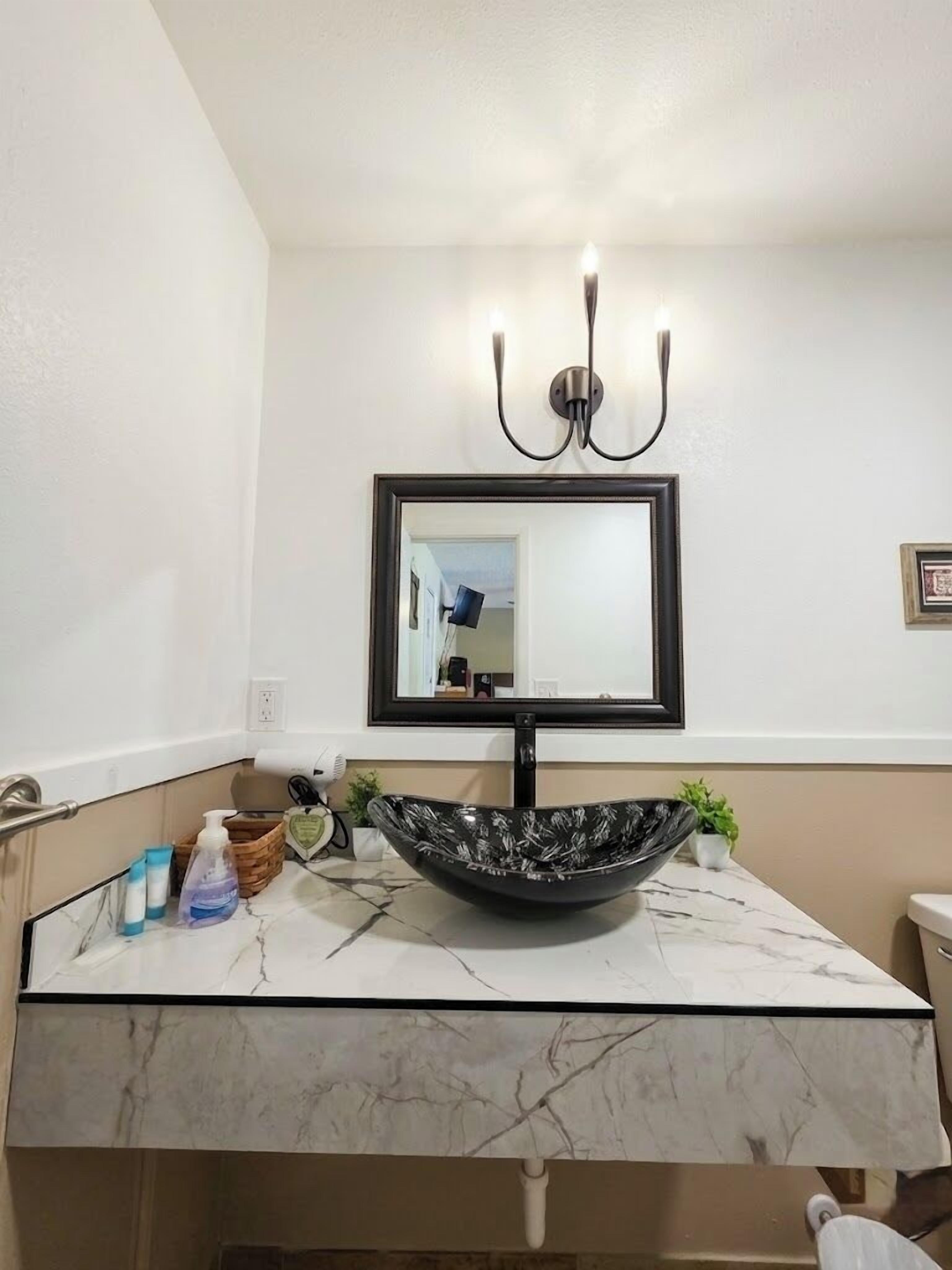 Un baño con lavamanos de mármol, un lavabo negro, un espejo con marco negro e iluminación empotrada en la pared.