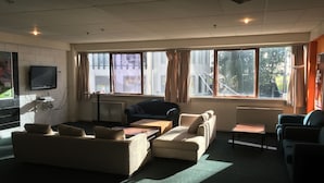 Uma sala de estar moderna com sofá, poltronas e mesa de centro. Há uma televisão na parede e grandes janelas com cortinas.