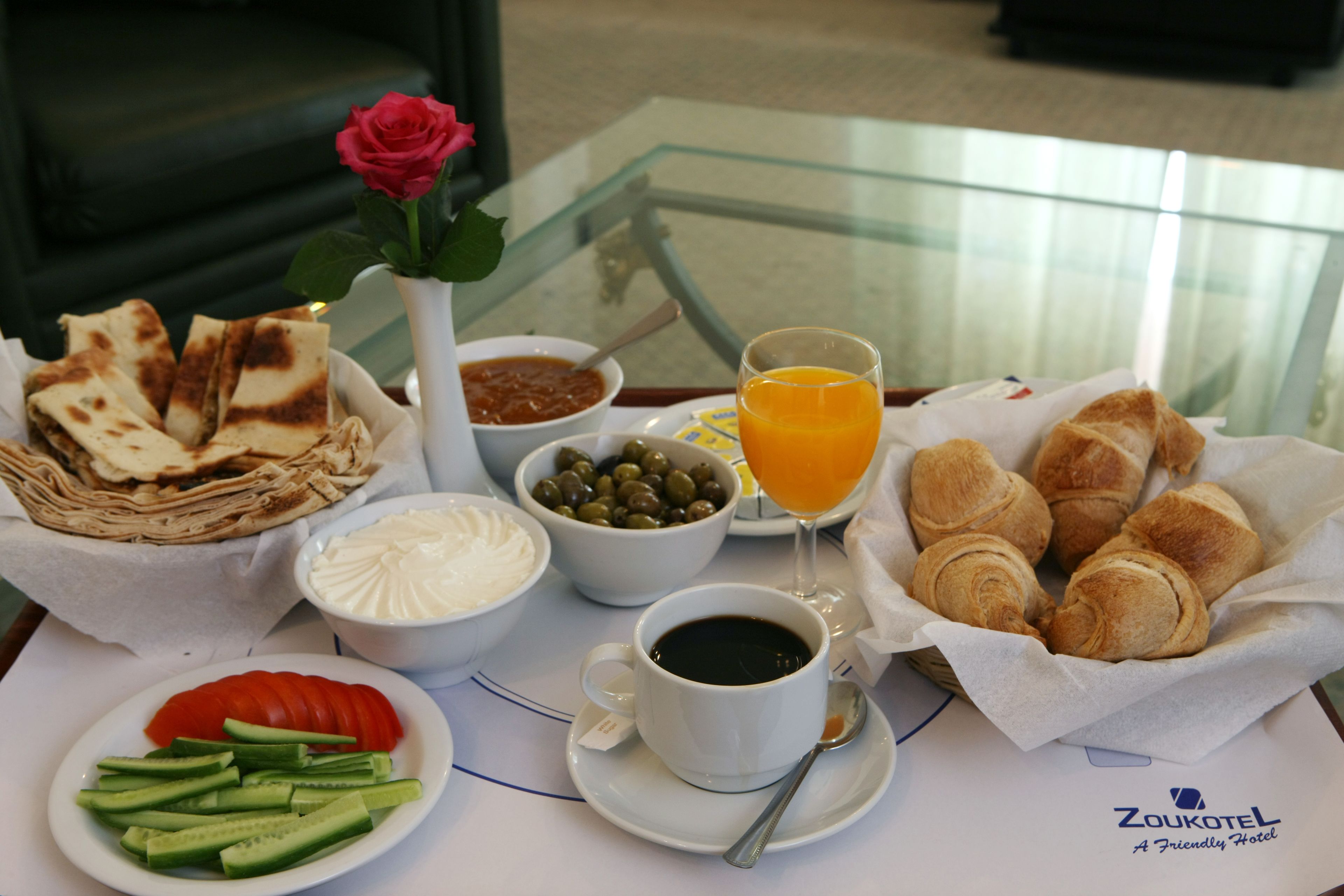 A breakfast table with croissants, a glass of orange juice, a cup of coffee, a bowl of olives, a bowl of yogurt, a plate of sliced cucumbers, and a rose in a vase.