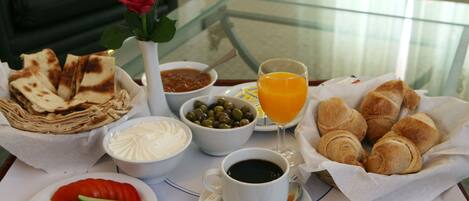 A breakfast table with croissants, a glass of orange juice, a cup of coffee, a bowl of olives, a bowl of yogurt, a plate of sliced cucumbers, and a rose in a vase.