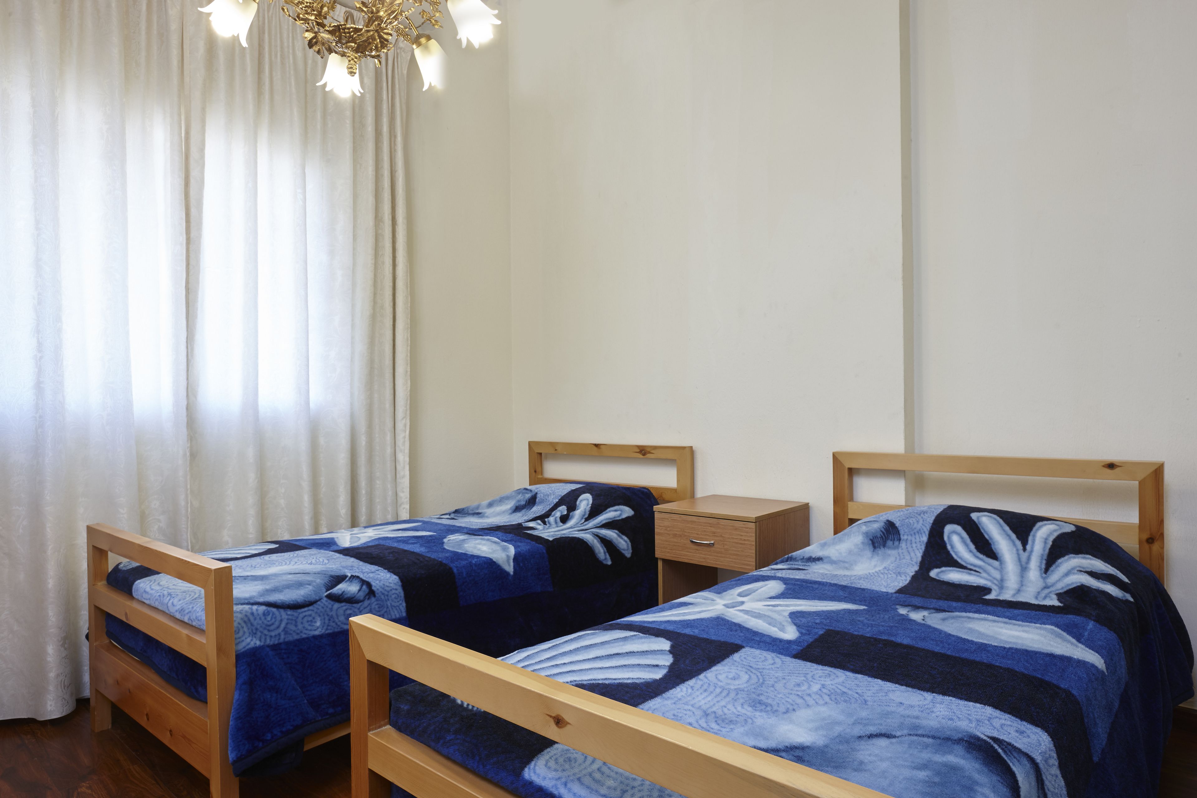 Two wooden beds with matching blue and white bedding, a wooden nightstand, and a window with sheer curtains.