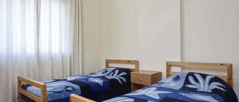 Two wooden beds with matching blue and white bedding, a wooden nightstand, and a window with sheer curtains.