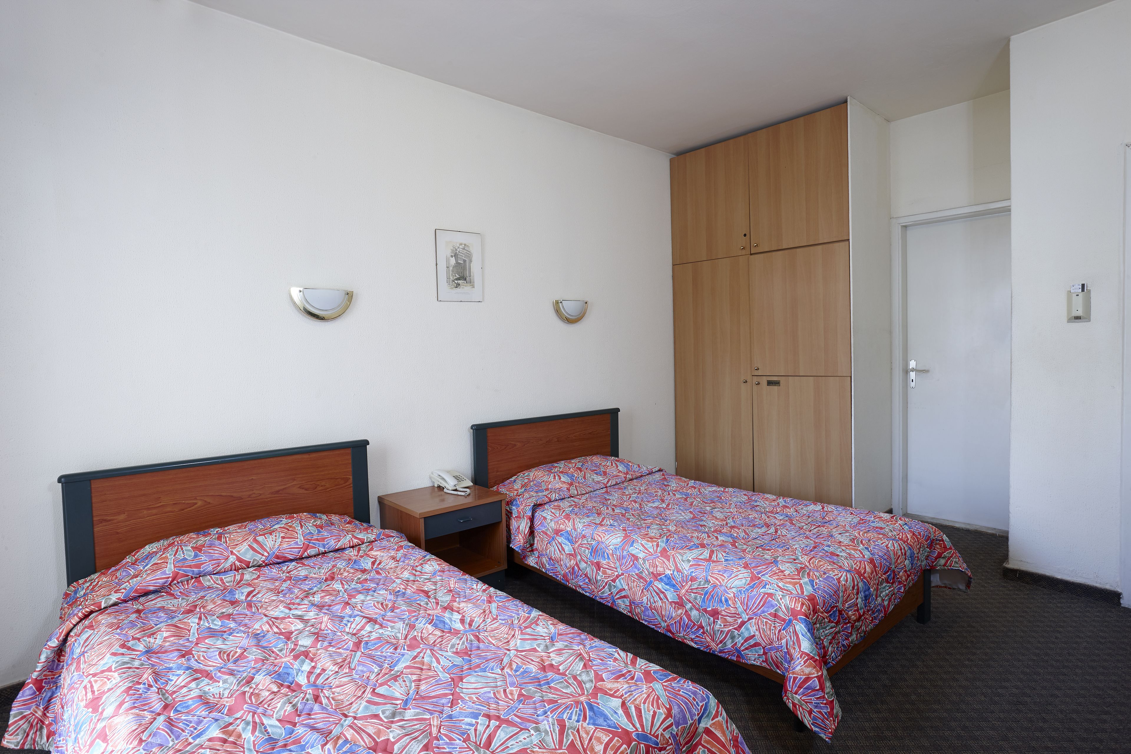 A hotel room with two single beds, a wooden wardrobe, a bedside table with a phone, and a door.