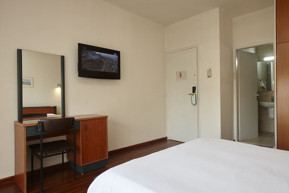 Una habitación de hotel con una cama, un escritorio con una silla, un televisor montado en la pared y una puerta que da al baño.