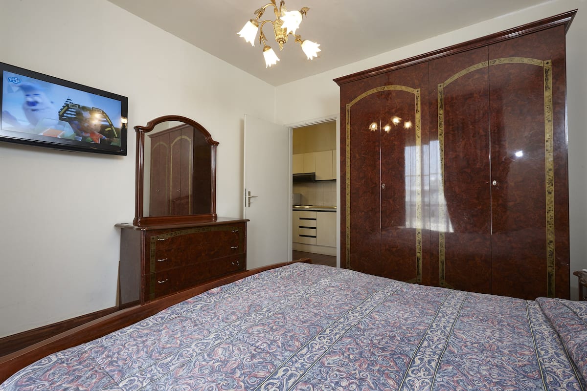 Un dormitorio con una cama grande, un armario de madera, un televisor en la pared y una cocina al fondo.