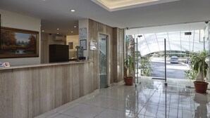 A hotel reception area with a wooden reception desk, a painting, a digital screen, and a glass door leading to a parking area with a view of a greenhouse. - Zoukotel Hotel (Jounieh)