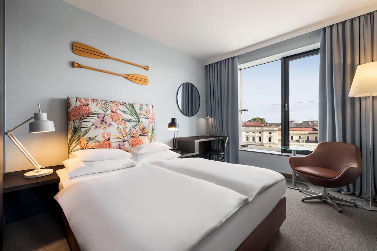A hotel room with a bed, a desk, a chair, a desk lamp, a painting, a round mirror, and a view of the city through the window.