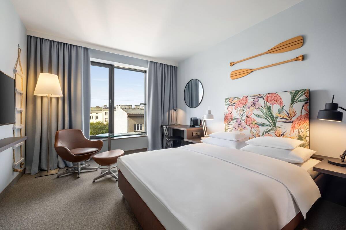A hotel room with a bed, a desk, a chair, a window with curtains, a painting, and two wooden paddles on the wall.