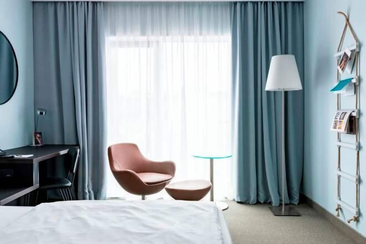 A hotel room with a bed, a desk, a chair, a small table, a lamp, and a window with curtains.