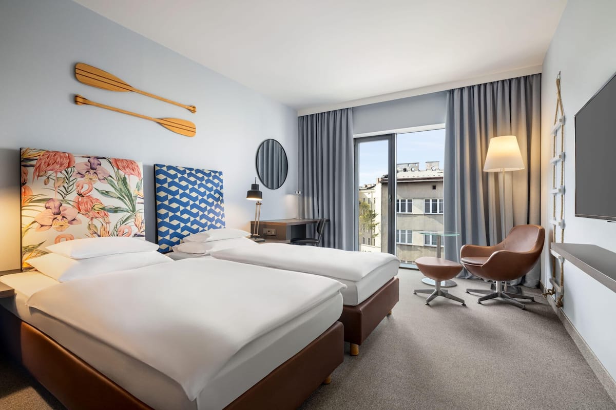 A hotel room with two beds, a desk, a chair, a TV, and a view of buildings through the window.