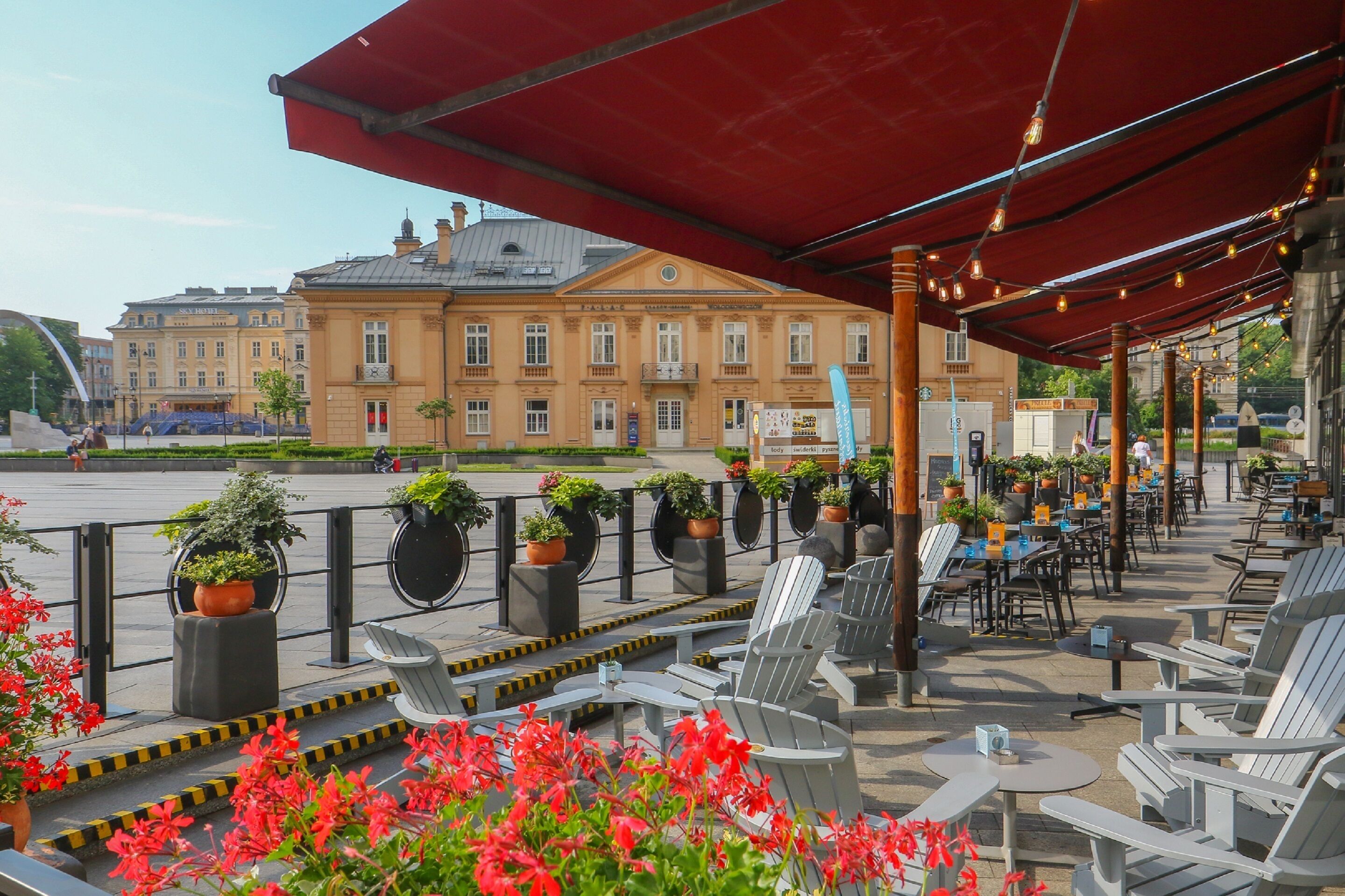 An outdoor cafe with tables and chairs, potted plants, and a red awning.