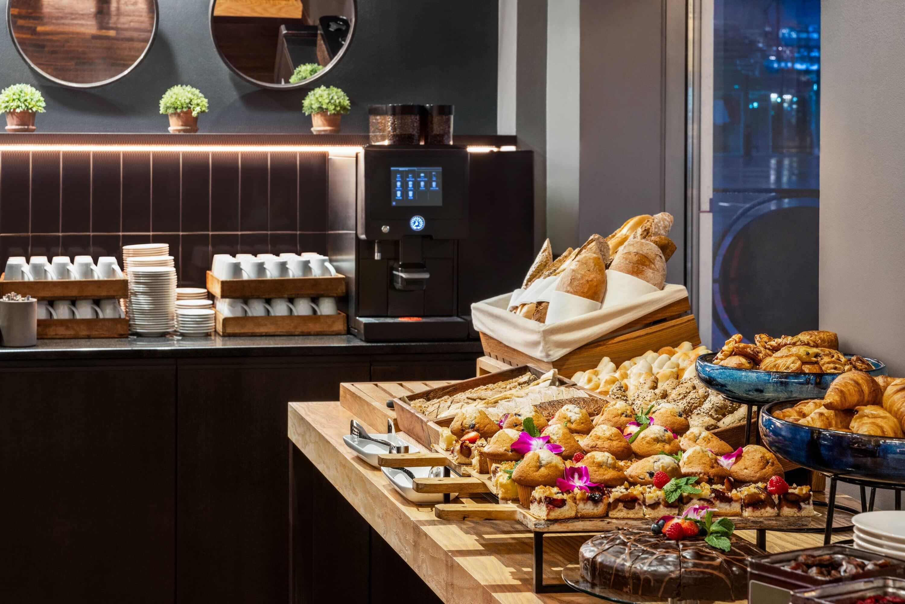 A buffet with various pastries, a coffee machine, and a view of the city at night.