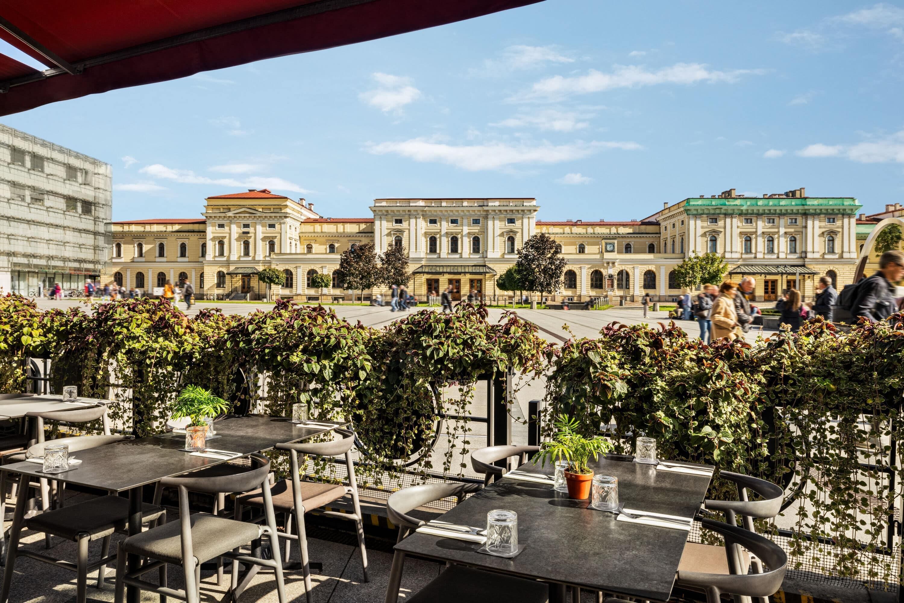 An outdoor café with tables and chairs, surrounded by lush greenery and historic buildings in the background.