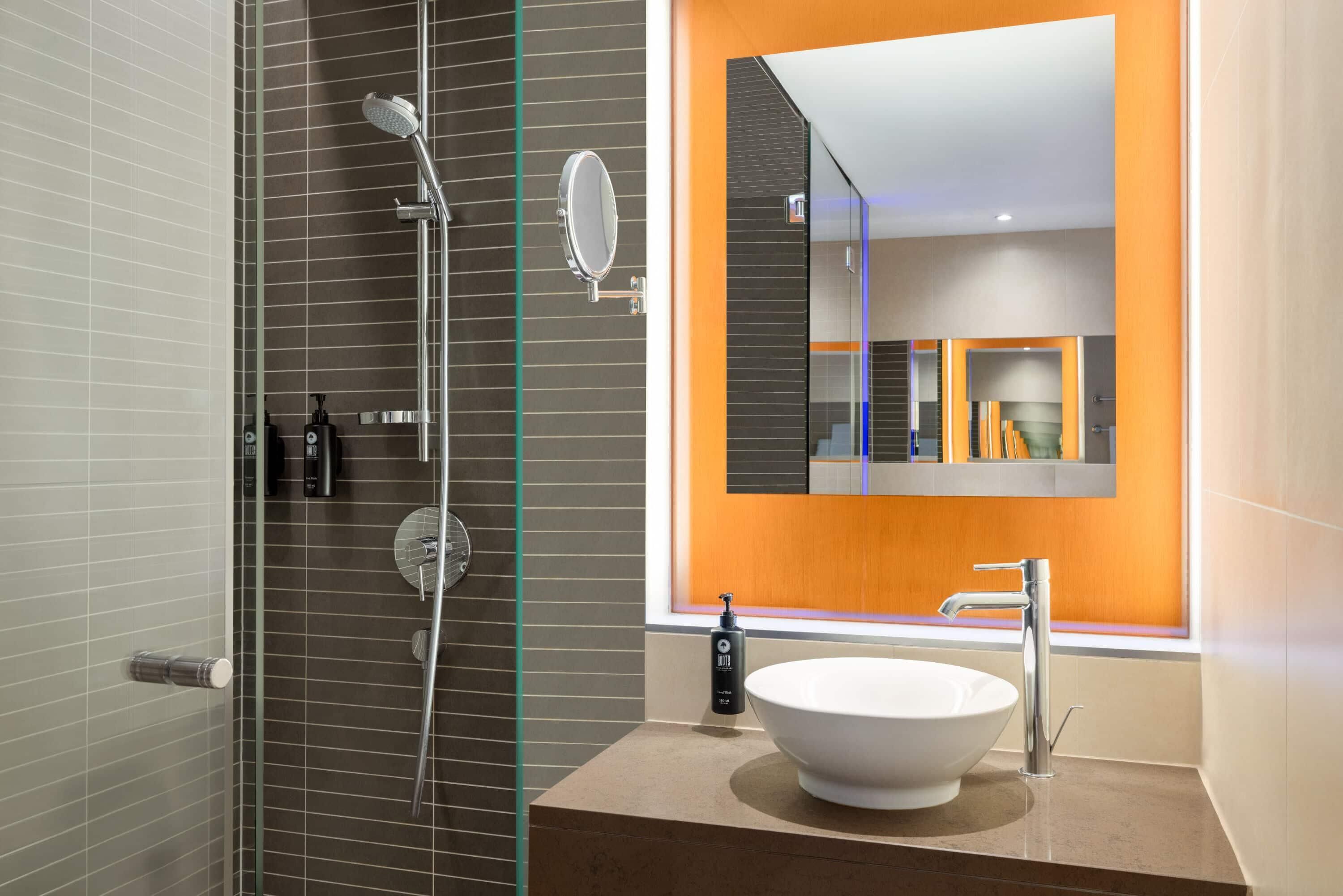 Un baño moderno con ducha envuelta en vidrio, un lavamanos ovalado grande y un espejo con una pared de acento naranja.