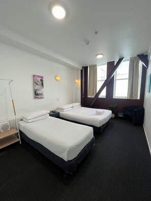 A hotel room with two single beds, a desk, a chair, a standing lamp, and a window with curtains.