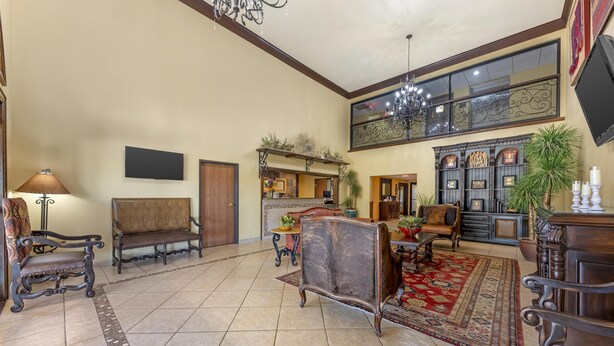 A spacious living room with a high ceiling, a chandelier, and a large window with decorative ironwork. There are several pieces of antique furniture, including a sofa and a chair, and a red patterned rug on the floor. - Best Western San Isidro Inn (Laredo)