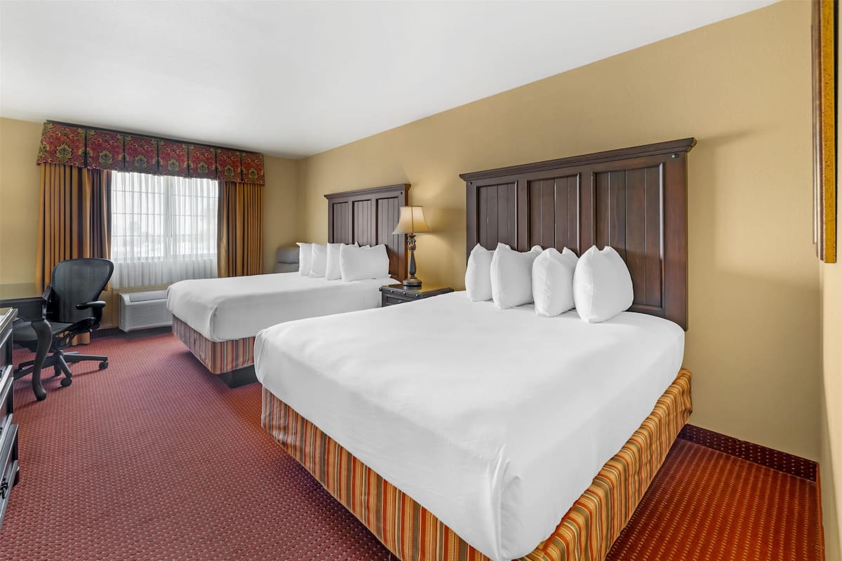 Quarto de hotel com duas camas, uma escrivaninha, uma cadeira e uma janela com cortinas.